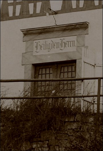 Ein historisches Gebäude mit einer Eingangstür, über der der Schriftzug "Heiligendamm" in geschwungener Schrift zu sehen ist. Die Tür ist von einem steinernen Rahmen umgeben und von einer einfachen Geländerstruktur im Vordergrund umgeben.