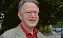 Ein älterer Mann mit Brille und grauen Haaren lächelt in die Kamera. Er trägt ein rotes Hemd und einen hellen Blazer. Im Hintergrund sind Bäume und eine Straße sichtbar.