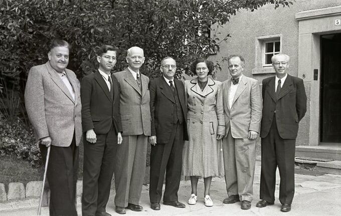 Eine Gruppe von acht Personen steht in einer Reihe vor einem Gebäude. Sie tragen formelle Kleidung und lächeln in die Kamera. Im Hintergrund sind Bäume und ein Fenster sichtbar.
