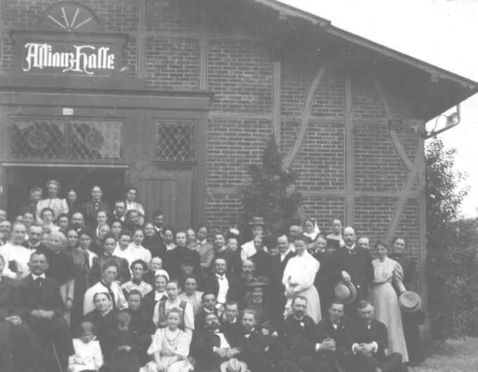 Eine große Gruppe von Menschen steht vor einem Gebäude mit der Aufschrift "Allianzhalle". Die Personen sind in historischen Kleidern gekleidet und lächeln in die Kamera. Es gibt Männer, Frauen und Kinder, die sich in mehreren Reihen aufstellen.