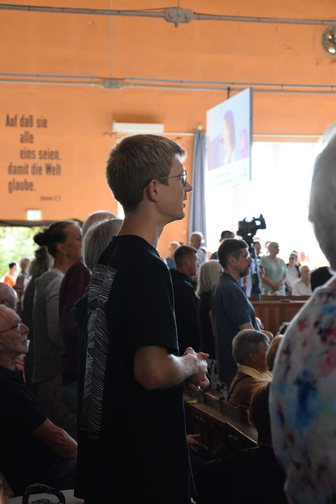 Ein junger Mann steht in einer Menschenmenge während einer Veranstaltung. Im Hintergrund sind weitere Personen zu sehen, und ein Bildschirm zeigt eine Präsentation. Die Atmosphäre wirkt aufmerksam und konzentriert.