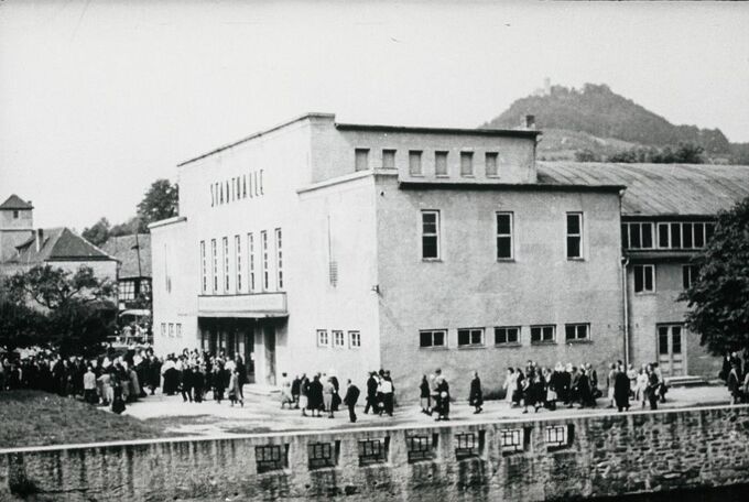 Eine historische Schwarz-Weiß-Aufnahme eines Gebäudes mit der Aufschrift "Säulenhalle". Vor dem Gebäude versammelt sich eine große Menschenmenge. Im Hintergrund ist ein Hügel mit einer Burg zu sehen.