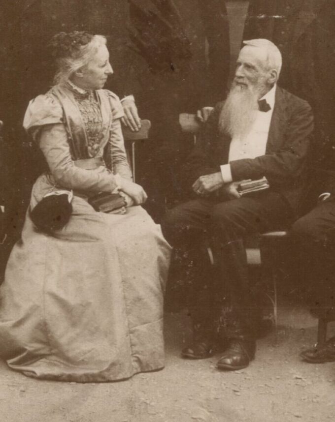 Ein älteres Paar sitzt sich gegenüber. Die Frau trägt ein elegantes, historisches Kleid und hat lange, lockige Haare. Der Mann hat einen langen weißen Bart und trägt einen Anzug. Beide scheinen ein Gespräch zu führen. Im Hintergrund sind unscharfe Silhouetten von weiteren Personen zu sehen.