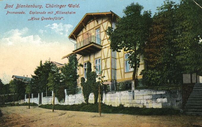 Eine historische Postkarte zeigt ein Gebäude mit Fachwerkarchitektur in Bad Blankenburg, Thüringer Wald. Umgeben von Bäumen und einer Promenade, ist das Haus mit dem Namen „Haus Gnadenfülle“ beschriftet. Der Himmel ist blau mit einigen Wolken.