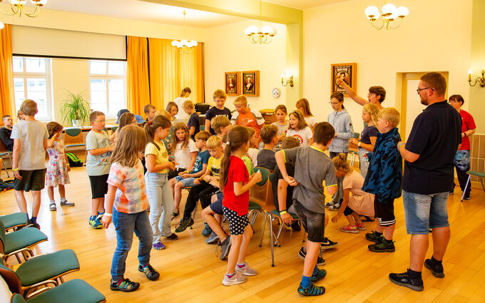 Eine Gruppe von Kindern und Jugendlichen versammelt sich in einem hellen Raum mit Holzfußboden. Einige stehen, andere sitzen auf Stühlen. Es herrscht eine fröhliche Atmosphäre, während die Kinder miteinander interagieren und spielen. Im Hintergrund sind Fenster und einige Wandbilder sichtbar.