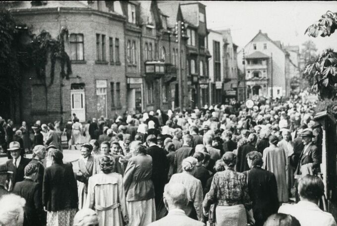 Eine große Menschenmenge steht auf einer Straße, umgeben von historischen Gebäuden. Die Personen sind unterschiedlich gekleidet und scheinen an einem Ereignis oder einer Versammlung teilzunehmen. Die Atmosphäre wirkt geschäftig und lebhaft.
