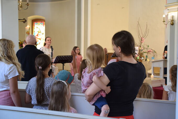 Eine Gruppe von Menschen in einer Kirche, die einem Auftritt zuschaut. Im Vordergrund stehen Frauen und Kinder, während eine junge Frau mit Mikrofon auf der Bühne spricht. Die Atmosphäre ist freundlich und gemeinschaftlich.