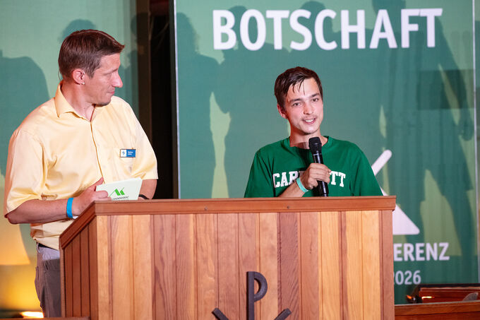 Ein junger Mann steht am Rednerpult und spricht ins Mikrofon, während ein Mann neben ihm aufmerksam zuhört. Im Hintergrund ist ein Banner mit der Aufschrift "BOTSCHAFT" zu sehen. Die Szene findet in einem festlichen Rahmen statt.