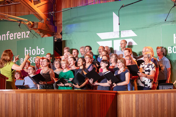 Ein gemischter Chor mit vielen Sängern steht auf einer Bühne und singt. Eine Dirigentin leitet den Chor. Im Hintergrund sind grüne Banner mit Texten zu sehen. Die Atmosphäre ist festlich und die Sänger sind konzentriert.