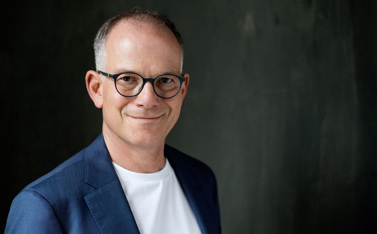 Ein lächelnder Mann mit Brille trägt einen blauen Blazer und ein weißes T-Shirt. Der Hintergrund ist dunkel und strukturiert.