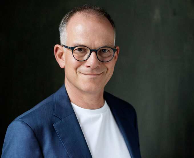 Ein lächelnder Mann mit Brille trägt einen blauen Blazer und ein weißes T-Shirt. Der Hintergrund ist dunkel und strukturiert.