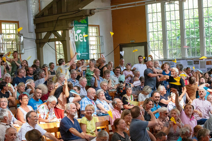 Eine große Menschenmenge sitzt in einem Saal und hebt gelbe Gegenstände in die Luft. Die Anwesenden wirken begeistert und sind in Gespräche vertieft. Im Hintergrund sind Fenster und eine grüne Banner mit dem Wort "bet" sichtbar.
