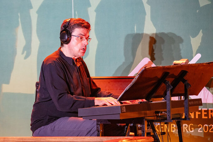 Ein Musiker sitzt an einem Keyboard und spielt. Er trägt Kopfhörer und schaut konzentriert auf die Noten. Im Hintergrund sind schemenhafte Figuren zu sehen, die eine Aufführung andeuten.