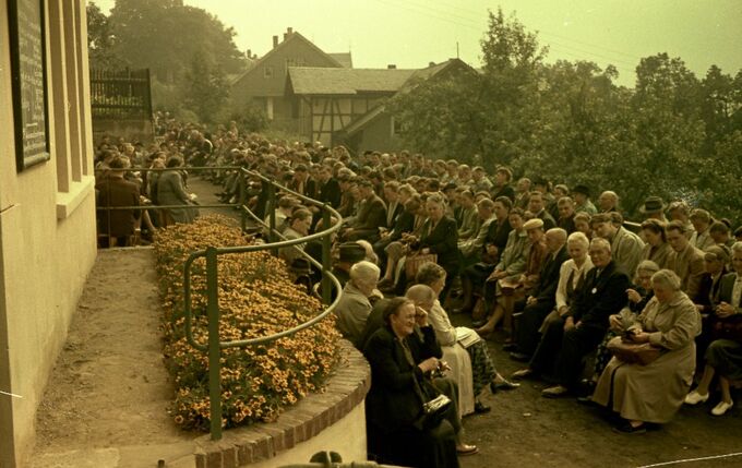 Eine große Menschenmenge sitzt auf einer geschwungenen Bank vor einem Gebäude, umgeben von Bäumen und Blumen. Die Anwesenden sind in formeller Kleidung und scheinen auf eine Veranstaltung zu warten. Im Hintergrund sind weitere Gebäude sichtbar.