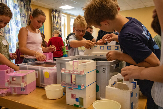 Kinder bauen mit Spielzeughäusern in einem Raum. Einige Kinder konzentrieren sich auf das Zusammenfügen von Bauteilen, während andere ihre Modelle präsentieren. Es gibt verschiedene Farben und Formen der Häuser, und die Atmosphäre ist kreativ und spielerisch.