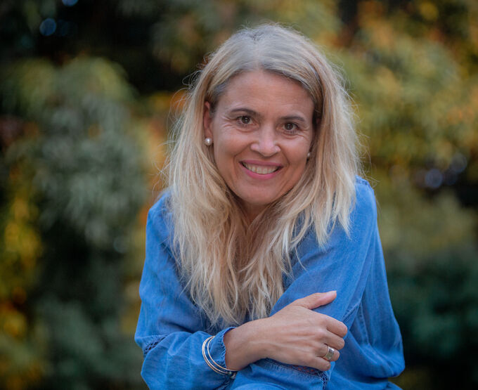 Eine lächelnde Frau mit langen, blonden Haaren trägt ein blaues Oberteil. Sie sitzt in einer natürlichen Umgebung mit Bäumen im Hintergrund und hat eine entspannte Haltung.