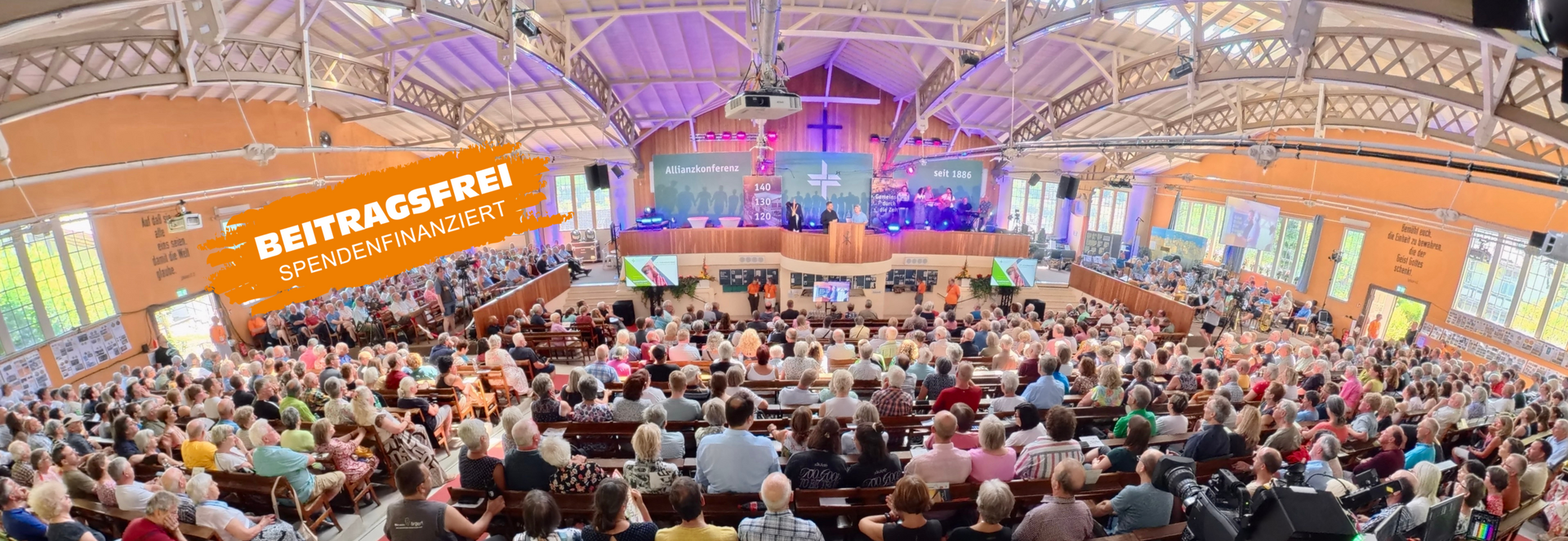 Eine große Menschenmenge sitzt in einem Kirchenraum. Die Wände sind hell, und es gibt eine Bühne mit Musikern. Ein Banner weist auf die Finanzierung durch Spenden hin.