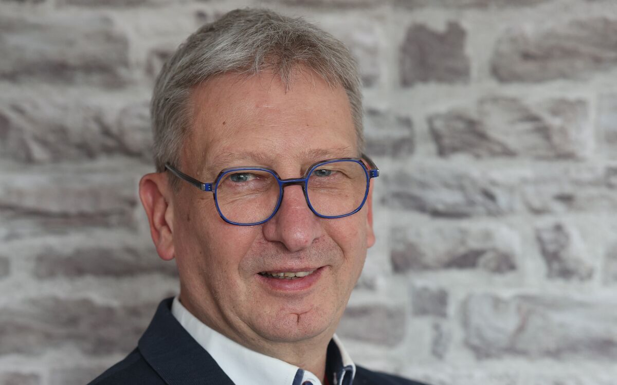 Ein Mann mit grauen Haaren und Brille lächelt in die Kamera. Er trägt einen dunklen Anzug und ein weißes Hemd mit einem Kragen. Im Hintergrund ist eine Steinmauer zu sehen.
