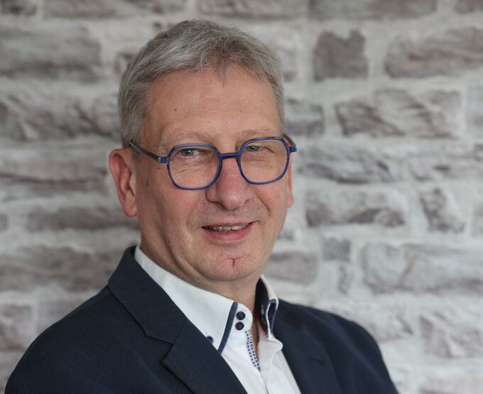 Ein Mann mit grauen Haaren und Brille lächelt in die Kamera. Er trägt einen dunklen Anzug und ein weißes Hemd mit einem Kragen. Im Hintergrund ist eine Steinmauer zu sehen.
