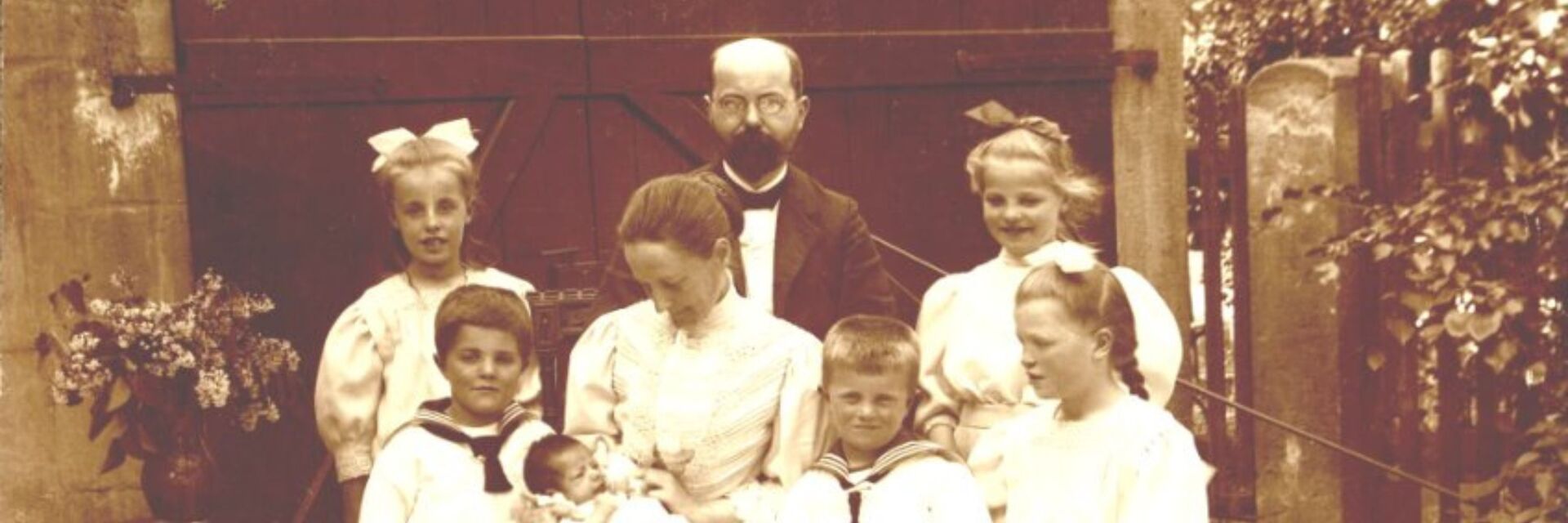 Eine historische Schwarz-Weiß-Fotografie zeigt eine Gruppe von sieben Kindern und einem Mann. Die Kinder tragen traditionelle Kleidung, einige mit Schleifen im Haar. Der Mann steht in der Mitte, umgeben von den Kindern, während eines von ihnen ein Baby hält. Im Hintergrund ist ein Tor zu sehen.
