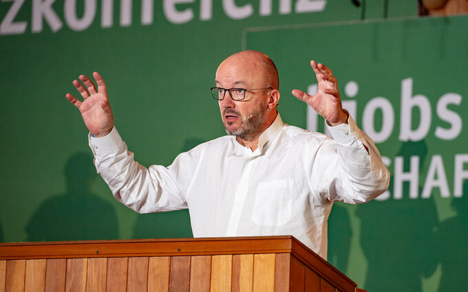 Ein Mann in einem weißen Hemd steht an einem Rednerpult und gestikuliert mit beiden Händen. Im Hintergrund sind grüne Wände mit Text zu sehen, der auf eine Konferenz hinweist. Der Mann wirkt engagiert und spricht wahrscheinlich zu einem Publikum.