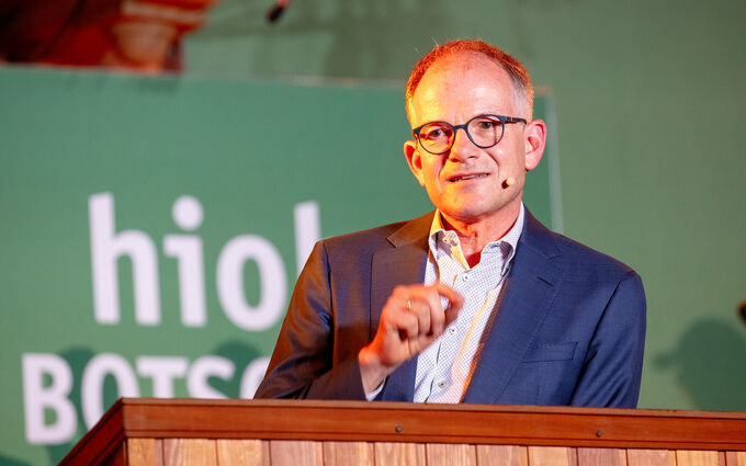 Ein Mann mit Brille spricht an einem Rednerpult. Er trägt einen Anzug und gestikuliert mit der Hand. Im Hintergrund ist ein grünes Banner mit dem Wort "hiol" und "BOTSCHAFTER" zu sehen.