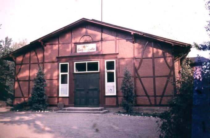 Ein historisches Gebäude mit rotem Ziegelfassade und Holzverzierungen. Es hat große Fenster und eine doppelte Eingangstür. Vor dem Gebäude stehen einige Bäume und Sträucher. Ein Schild mit der Aufschrift „Kulturstätte“ ist über dem Eingang angebracht.