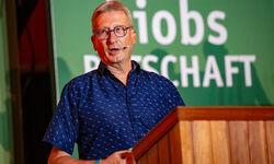 Ein Mann mit Brille und blauem Hemd spricht an einem Rednerpult. Im Hintergrund ist ein grünes Banner mit dem Text "Jobs" und "Wirtschaft" zu sehen. Die Szene vermittelt eine Atmosphäre einer Präsentation oder Konferenz. (Text KI generiert)