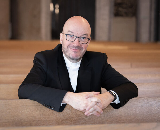 Ein Mann mit Brille und Glatze sitzt auf einer Kirchenbank. Er trägt einen schwarzen Anzug und ein weißes Hemd. Der Hintergrund zeigt eine helle, moderne Kirchenarchitektur. Der Mann lächelt und hat die Hände gefaltet.