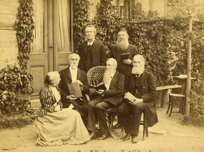 Sechs Personen in historischer Kleidung posieren im Freien vor einem Gebäude, umgeben von Pflanzen. Sie halten Bücher in den Händen und sitzen oder stehen in einer Gruppe. Die Szene vermittelt einen Eindruck von Gemeinschaft und Bildung.