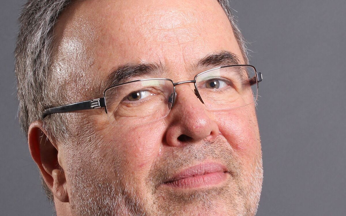 Ein Mann mit grauen Haaren und Brille schaut direkt in die Kamera. Er trägt ein schwarzes Hemd und hat einen leichten Bart. Der Hintergrund ist grau.