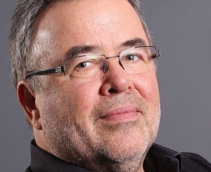 Ein Mann mit grauen Haaren und Brille schaut direkt in die Kamera. Er trägt ein schwarzes Hemd und hat einen leichten Bart. Der Hintergrund ist grau.