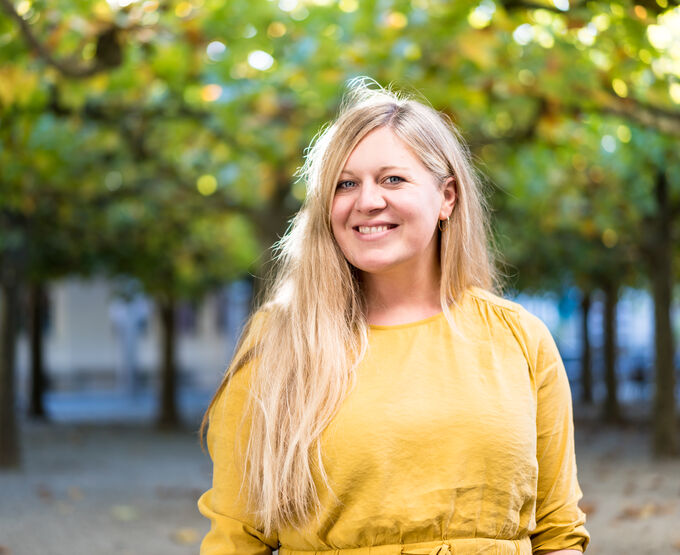 Eine lächelnde Frau mit langen, blonden Haaren trägt ein gelbes Kleid. Sie steht in einem Park mit Bäumen im Hintergrund, die grüne Blätter haben. Das Licht ist weich und schafft eine freundliche Atmosphäre.