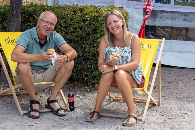 Zwei Personen sitzen entspannt in Liegestühlen im Freien. Sie genießen Snacks und eine Flasche Cola. Im Hintergrund sind Pflanzen und ein Zelt sichtbar. Die Atmosphäre wirkt freundlich und gemütlich.