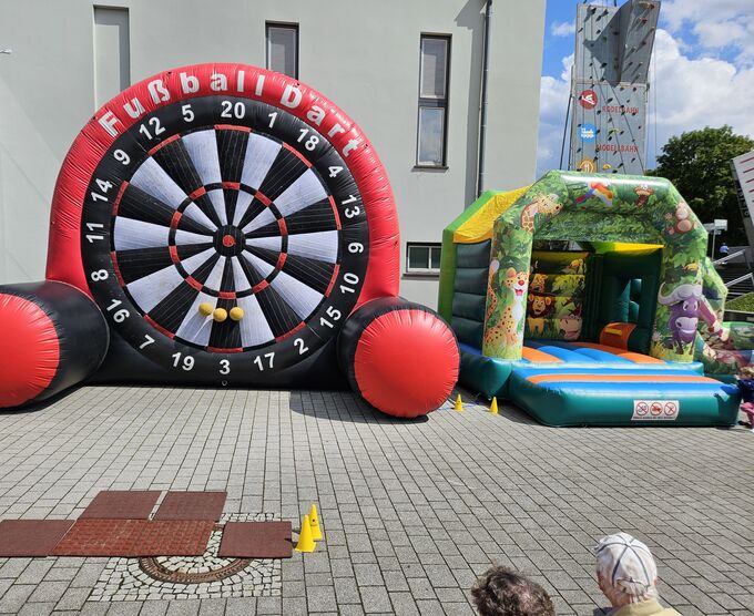 Ein aufblasbares Dartboard mit großen roten Rändern steht links, daneben ein aufblasbares Spielhaus mit buntem Design. Der Boden ist gepflastert, und im Vordergrund sind einige gelbe Kegel zu sehen. Der Himmel ist teilweise bewölkt.