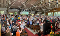 Eine große Menschenmenge steht in einem Gottesdienstraum, viele heben die Hände. Die Atmosphäre ist lebhaft und engagiert, mit Bänken und einer Bühne im Hintergrund. Helle Fenster lassen Tageslicht herein. (Text KI generiert)