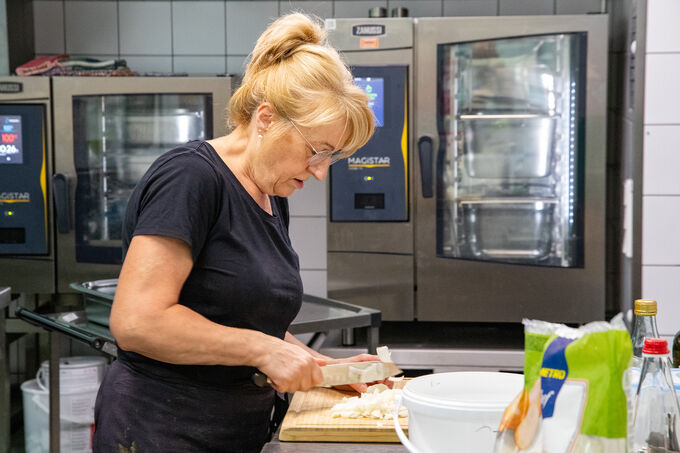 Eine Frau mit blonden Haaren schneidet Gemüse auf einem Holzbrett in einer modernen Küche. Im Hintergrund sind große Kochgeräte zu sehen. Die Frau trägt ein schwarzes T-Shirt und konzentriert sich auf ihre Arbeit.