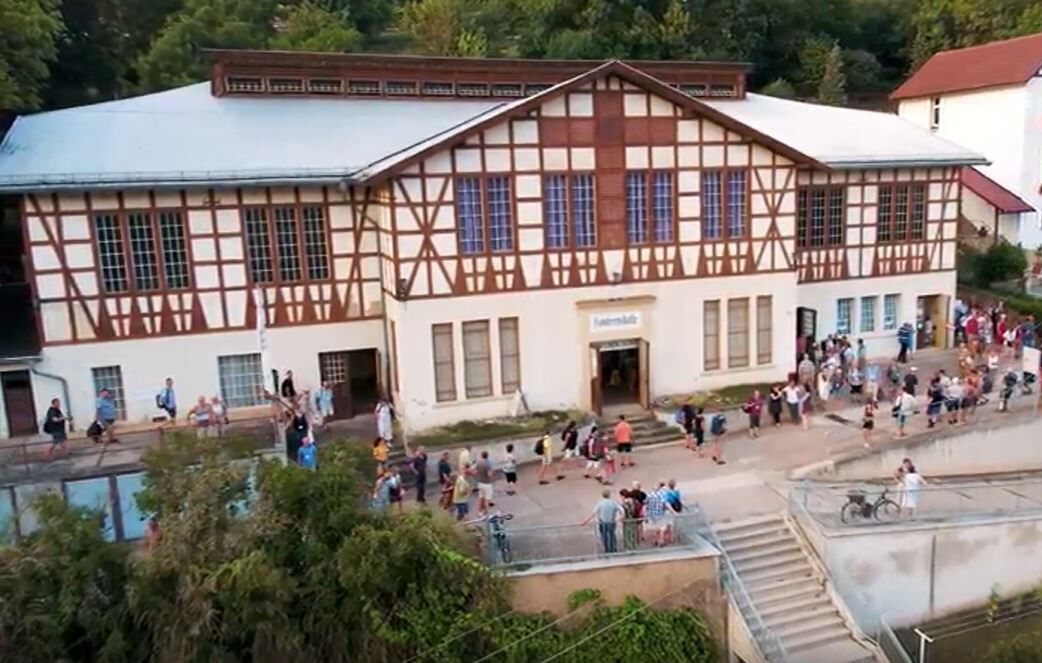 Ein historisches Gebäude mit Fachwerkarchitektur, umgeben von Bäumen. Vor dem Eingang stehen viele Menschen in einer Schlange. Einige Personen sind auf einem Gehweg und es gibt Fahrräder in der Nähe.