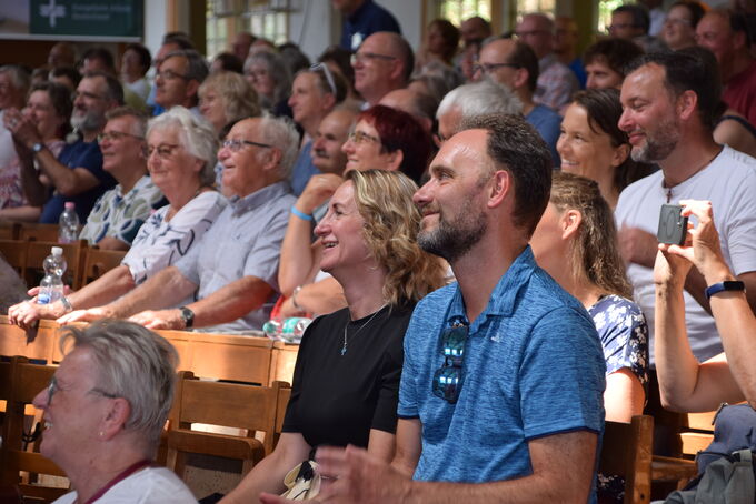 Eine große Menschenmenge sitzt in einem Saal und schaut lächelnd auf die Bühne. Einige Personen klatschen, während andere entspannt wirken. Die Zuschauer sind vielfältig und scheinen an einer Veranstaltung teilzunehmen.