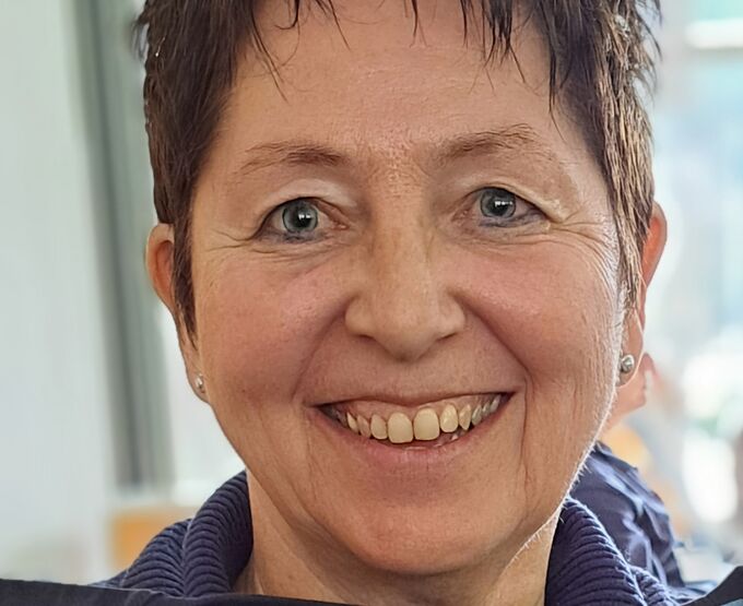 Eine lächelnde Frau mit kurzem, frisiertem Haar und blauen Augen. Sie trägt ein Oberteil in Blau und hat Ohrringe. Der Hintergrund ist unscharf, mit hellen Farben.