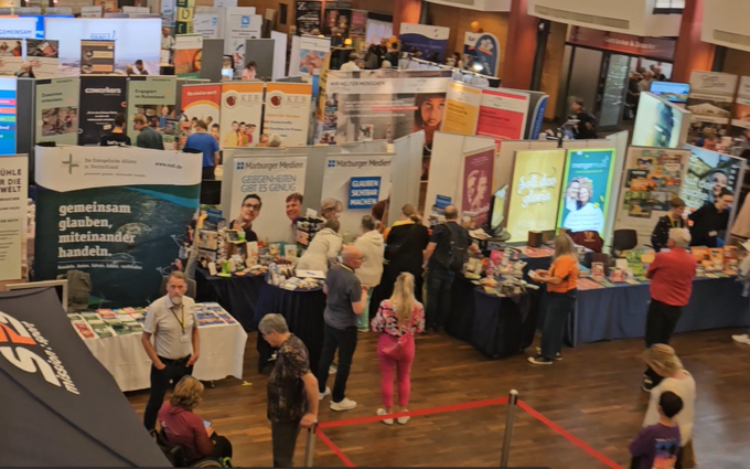 Eine Luftaufnahme einer Messehalle mit zahlreichen Ständen und Besuchern. Verschiedene Informationsstände sind zu sehen, die Produkte und Dienstleistungen präsentieren. Menschen bewegen sich zwischen den Ständen, während einige Gespräche führen. Ein großes Zelt mit dem Logo "SRS" ist im Vordergrund sichtbar.