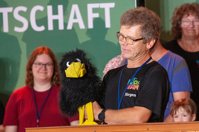 Ein Mann hält eine schwarze Handpuppe in Form eines Vogels und spricht mit ihr. Im Hintergrund sind mehrere Personen zu sehen, die aufmerksam zuschauen. Der Mann trägt ein T-Shirt mit der Aufschrift "Kids Team".