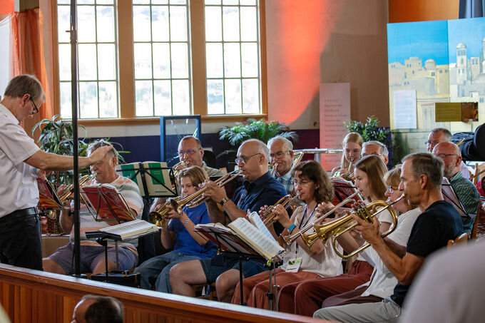 Eine Gruppe von Musikern sitzt in einem Raum und spielt Blasinstrumente. Ein Dirigent steht vorne und leitet die Aufführung. Im Hintergrund sind Fenster und Pflanzen zu sehen. Die Musiker sind konzentriert und haben Notenblätter vor sich.